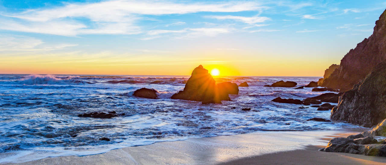 Vibrant sunset over a rocky shoreline, with waves gently washing onto a sandy beach under a partly cloudy sky.