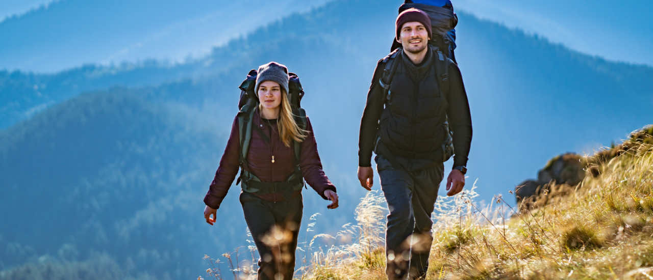 A couple wearing backpacks and outdoor gear hiking up a sunlit mountain trail, with layered blue ridges stretching into the distance, symbolizing a shared journey and partnership moving forward.