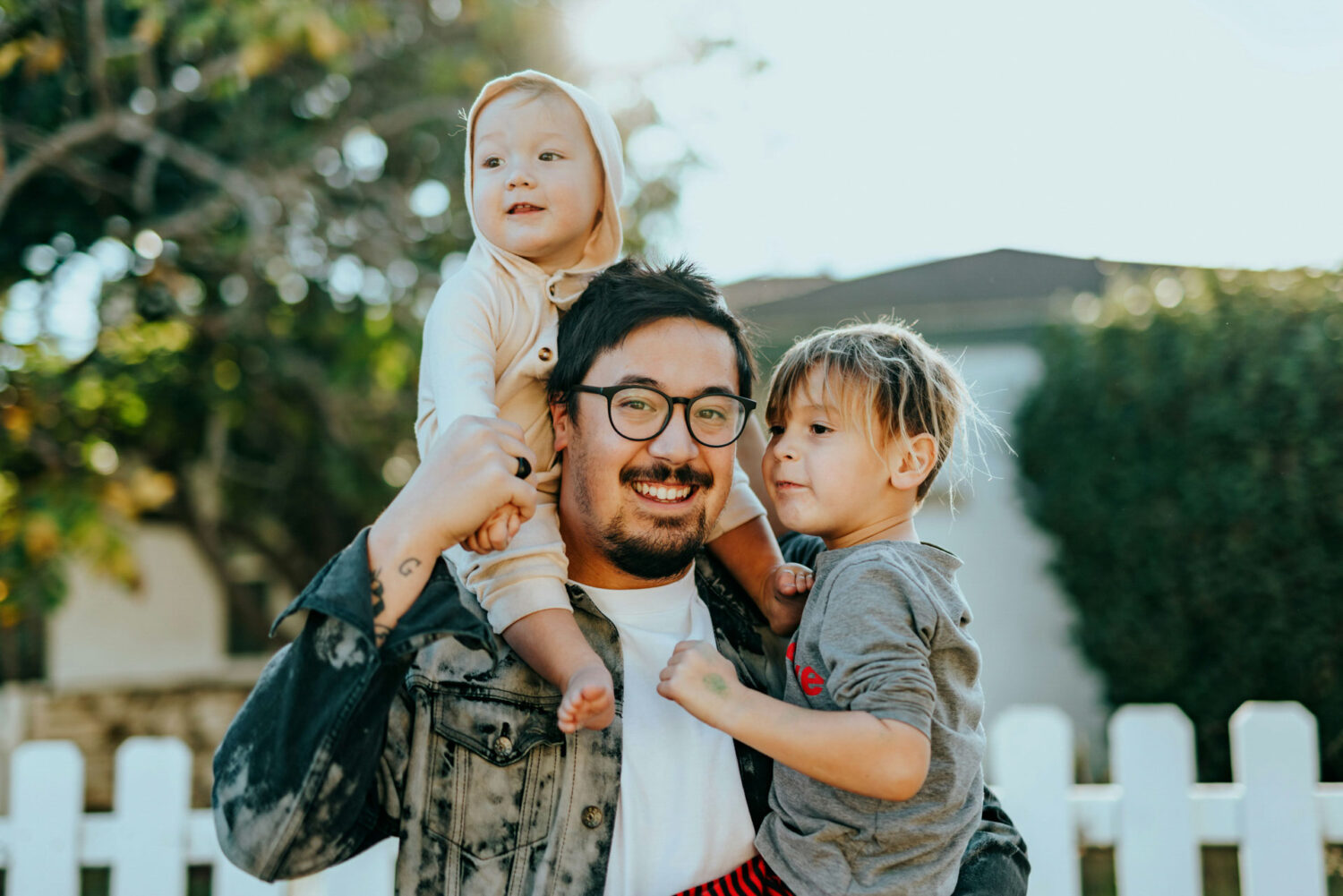 A smiling father holding two children on his shoulders in a bright outdoor setting.