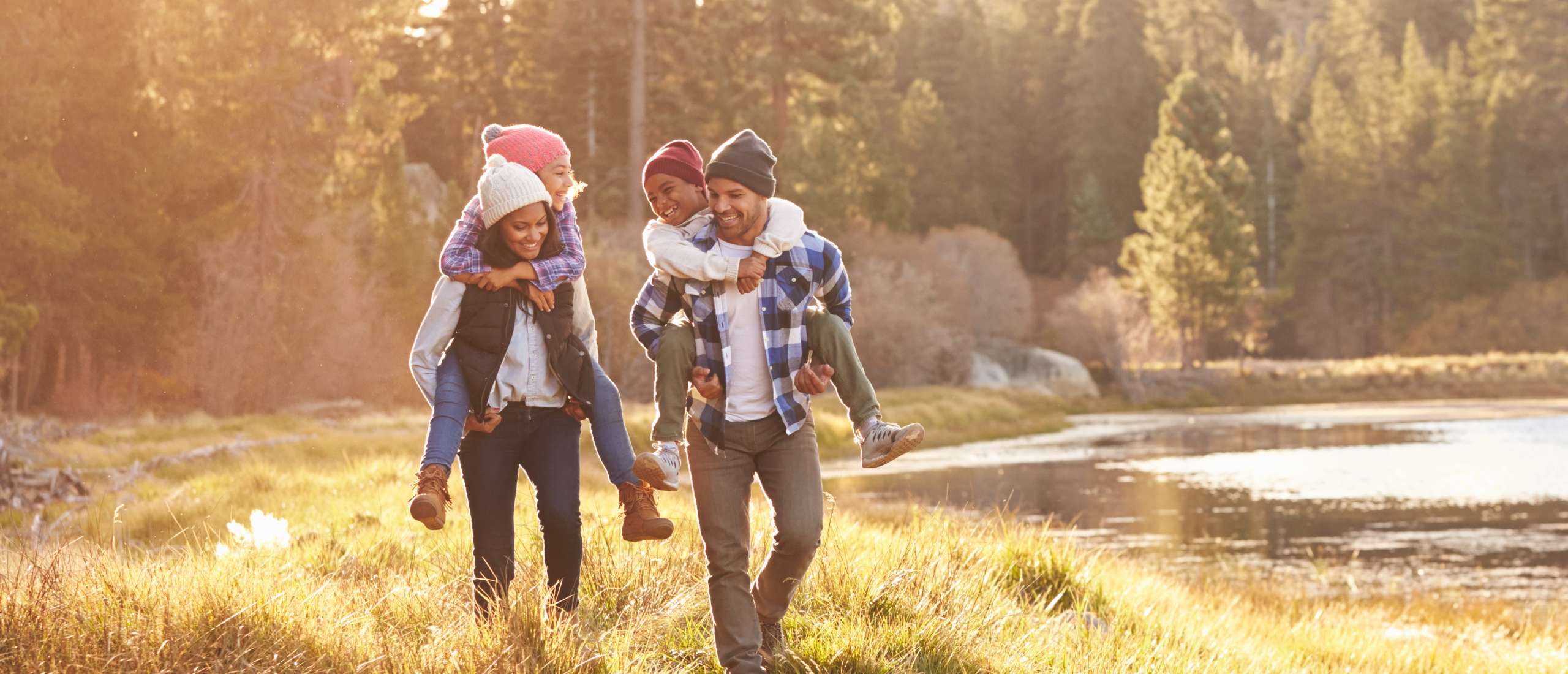 Parents giving piggyback rides to children wearing winter hats in golden sunlight near lake with mountain backdrop