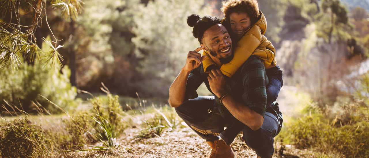 A smiling father crouches on a woodland trail while his young child, wearing a yellow jacket, rides piggyback among sun-dappled trees.