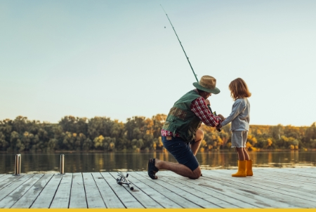 Father kneeling beside young daughter on wooden dock with fishing rod, teaching her to fish on calm lake at sunset