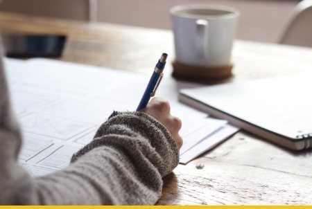 Hand in knit sweater holding pen while writing on paper with coffee cup and smartphone on desk