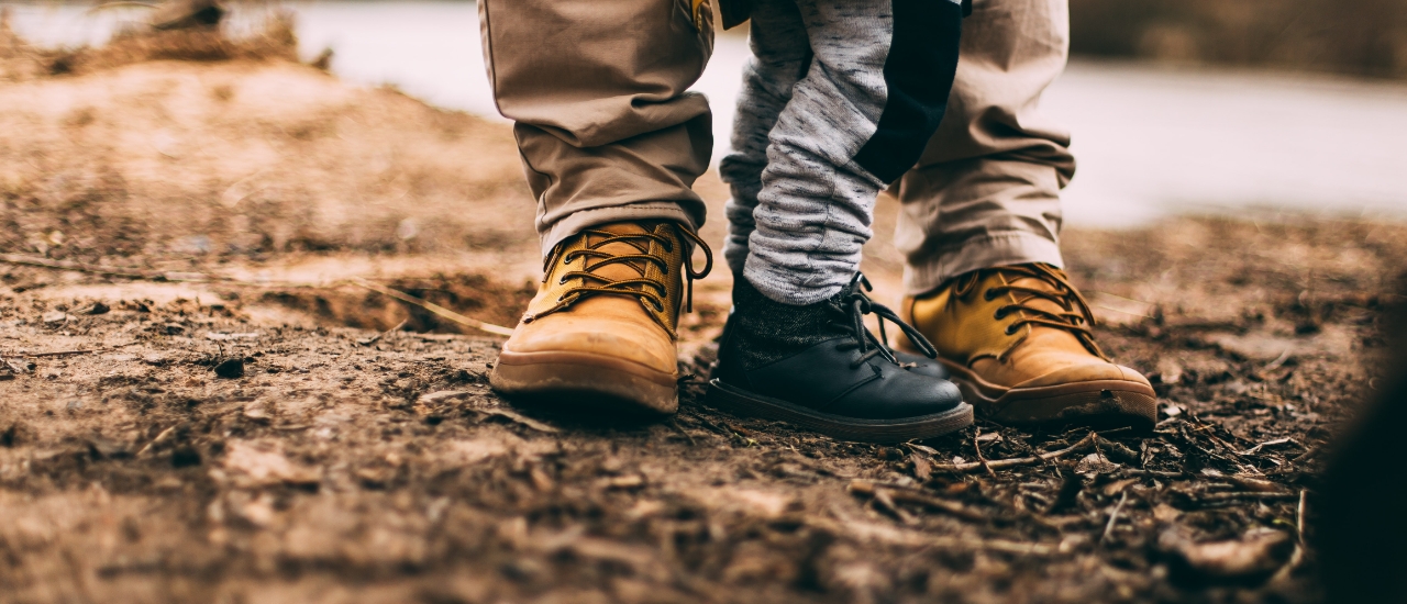 Close-up of two pairs of hiking boots on dirt trail - tan and black leather boots with laces and wool socks