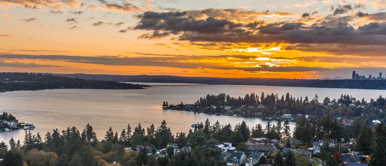 Aerial view of lakeside residential community with homes nestled among evergreen trees during golden sunset with dramatic cloudy sky