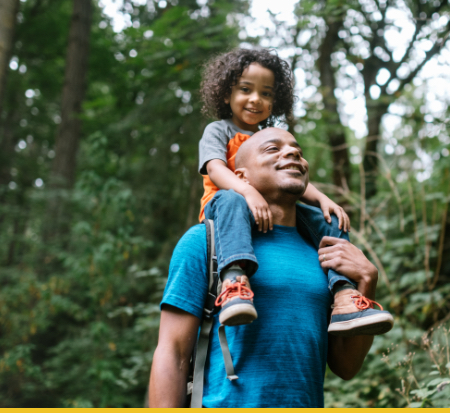 A father carrying his son on his shoulders while walking through a lush green forest.