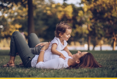 A pregnant mother lying on the grass with her smiling young child playing on her stomach outdoors.