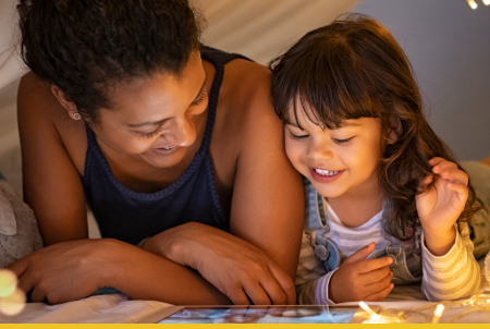 A smiling woman and a young girl enjoying time together while looking at a tablet in a cozy setting.