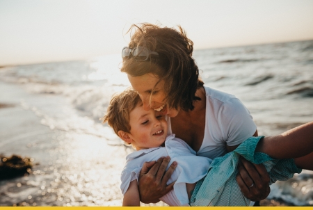 Mother and young son embracing on beach during golden hour sunset with ocean waves in background