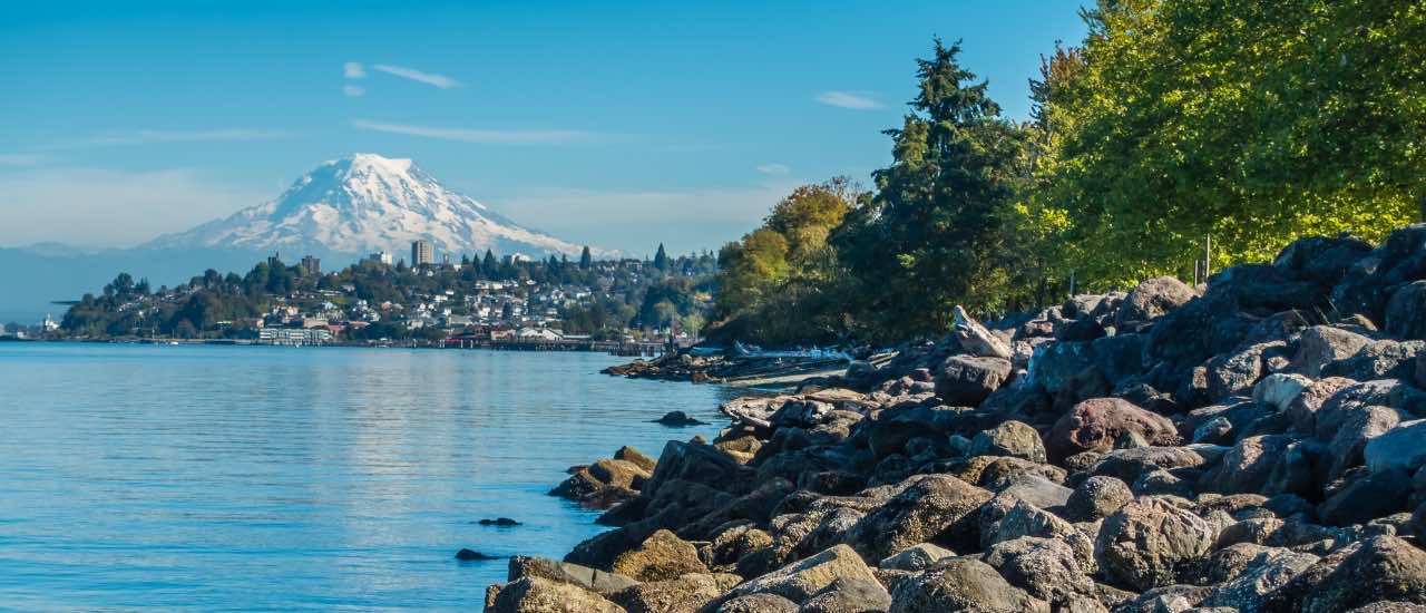 View of Mount Rainier rising beyond a rocky shoreline and tree-lined waterfront in Seattle, with calm blue water in the foreground.