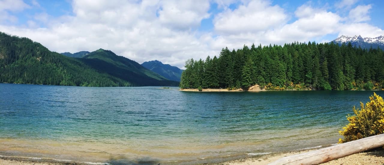 Clear mountain lake surrounded by dense forest with snow-capped peaks and cloudy sky