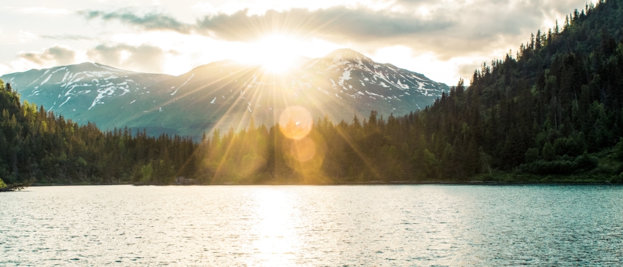 Sunrise casting rays over a forested lake with snow-capped mountains