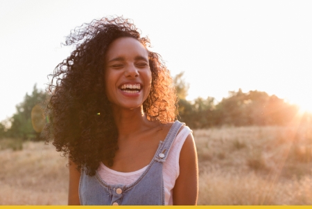 A young woman with curly hair smiling in a sunlit outdoor setting.