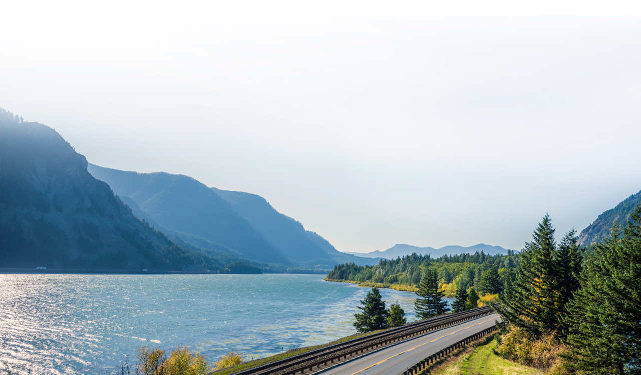 A winding highway and adjacent railroad tracks running alongside a shimmering mountain lake, flanked by evergreen forests and layered blue ridges under a pale sky.