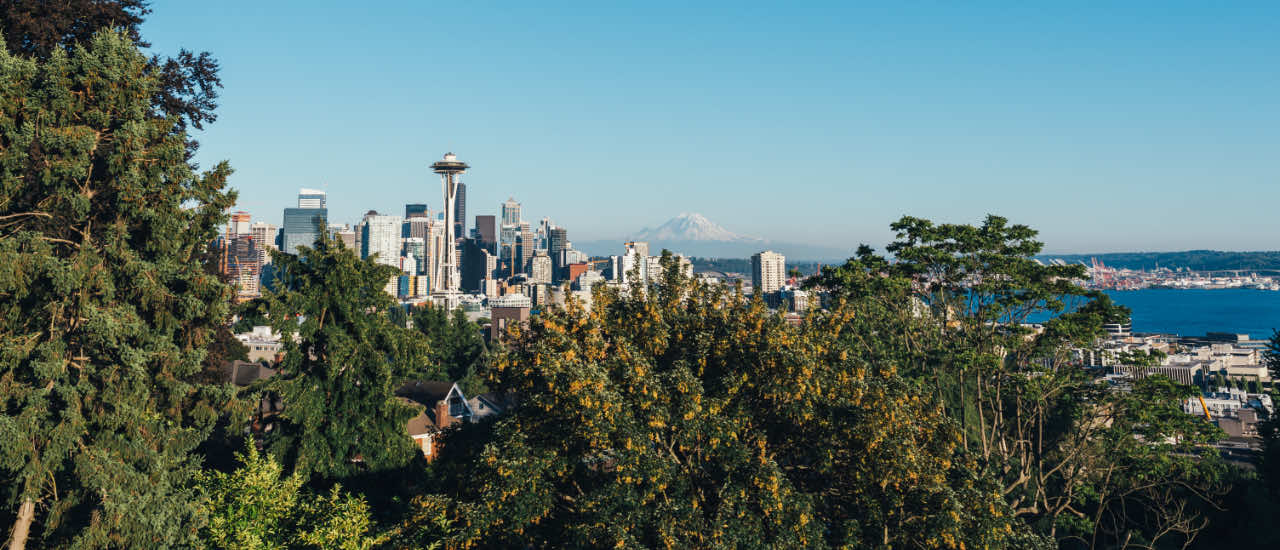 Panoramic view of Seattle’s skyline from Kerry Park, featuring the Space Needle centered among downtown skyscrapers, lush trees in the foreground, Elliott Bay to the right, and Mount Rainier rising in the background under a clear blue sky.