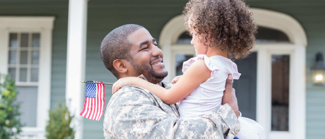 Uniformed service member smiling as he embraces his young daughter in front of their home, with the child holding a small American flag.