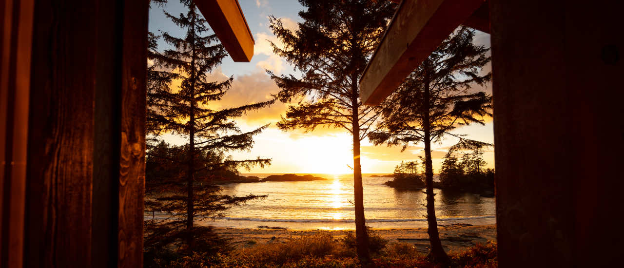 Golden sunset viewed through the open doorway of a wooden cabin, with silhouetted pine trees and small rocky islands on a calm ocean horizon.