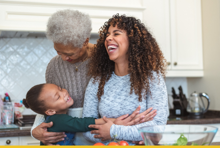 Three generations of women laughing together in bright white kitchen - grandmother, mother with curly hair, and young child
