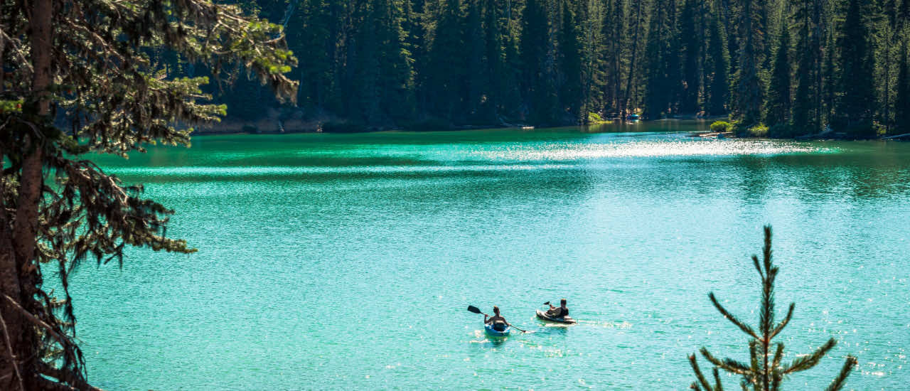 Two people kayaking across a calm, turquoise mountain lake bordered by dense pine forest, evoking a sense of tranquility and fresh beginnings.