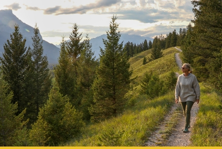A woman walking on a scenic trail surrounded by lush greenery and mountains in the background during sunset.