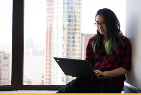 Woman with glasses working on laptop by large window overlooking city buildings in modern office space
