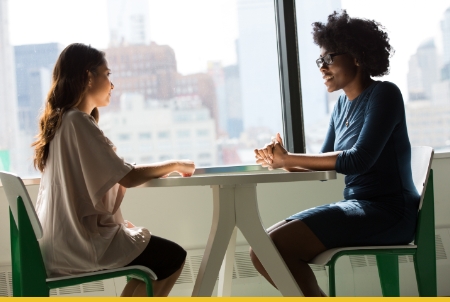 Two women engaged in a conversation at a table with a city skyline in the background.