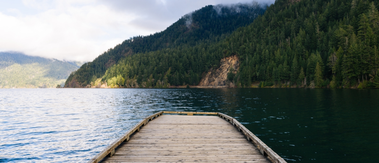 Wooden dock extending into calm mountain lake surrounded by forested hills and cloudy sky